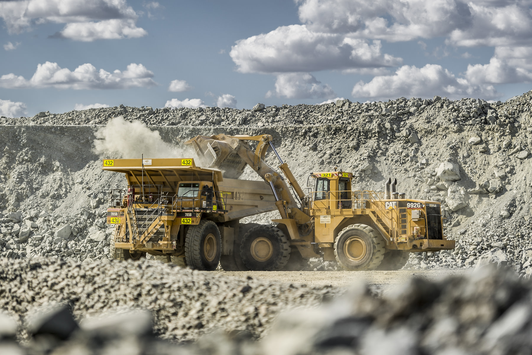 A dramatic shot of heavy machinery at a local quarry, featuring a front-end loader unloading onto a mining truck under a clear, sunlit sky.