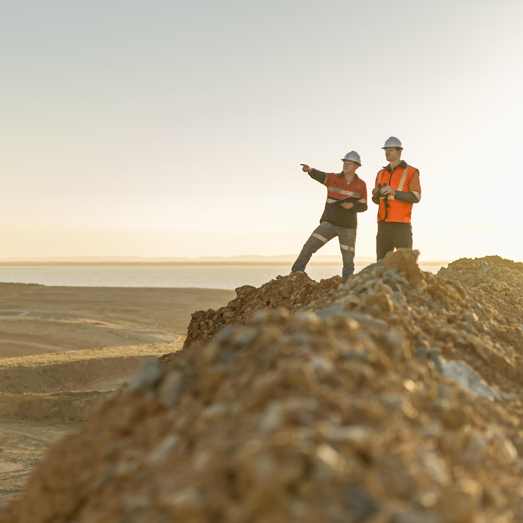 Industrial photograph of two engineers wearing safety gear and standing on a mound of dirt at a mine site during sunrise, with one pointing toward the horizon.